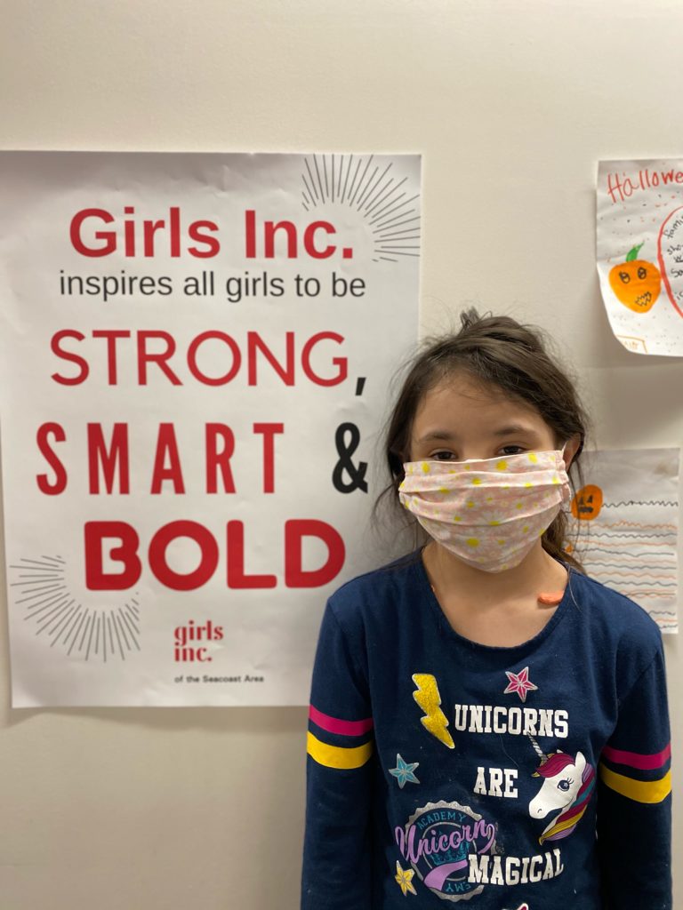 Image of a girl wearing a face mask, standing in front of a Girls Inc. poster that says "Strong, Smart & Bold"