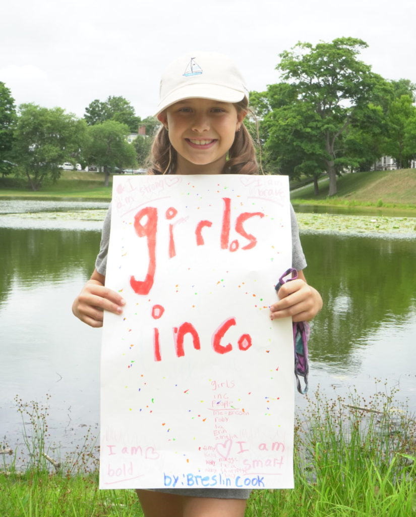 Young girl standing outside smiling, holding poster that reads Girls Inc.