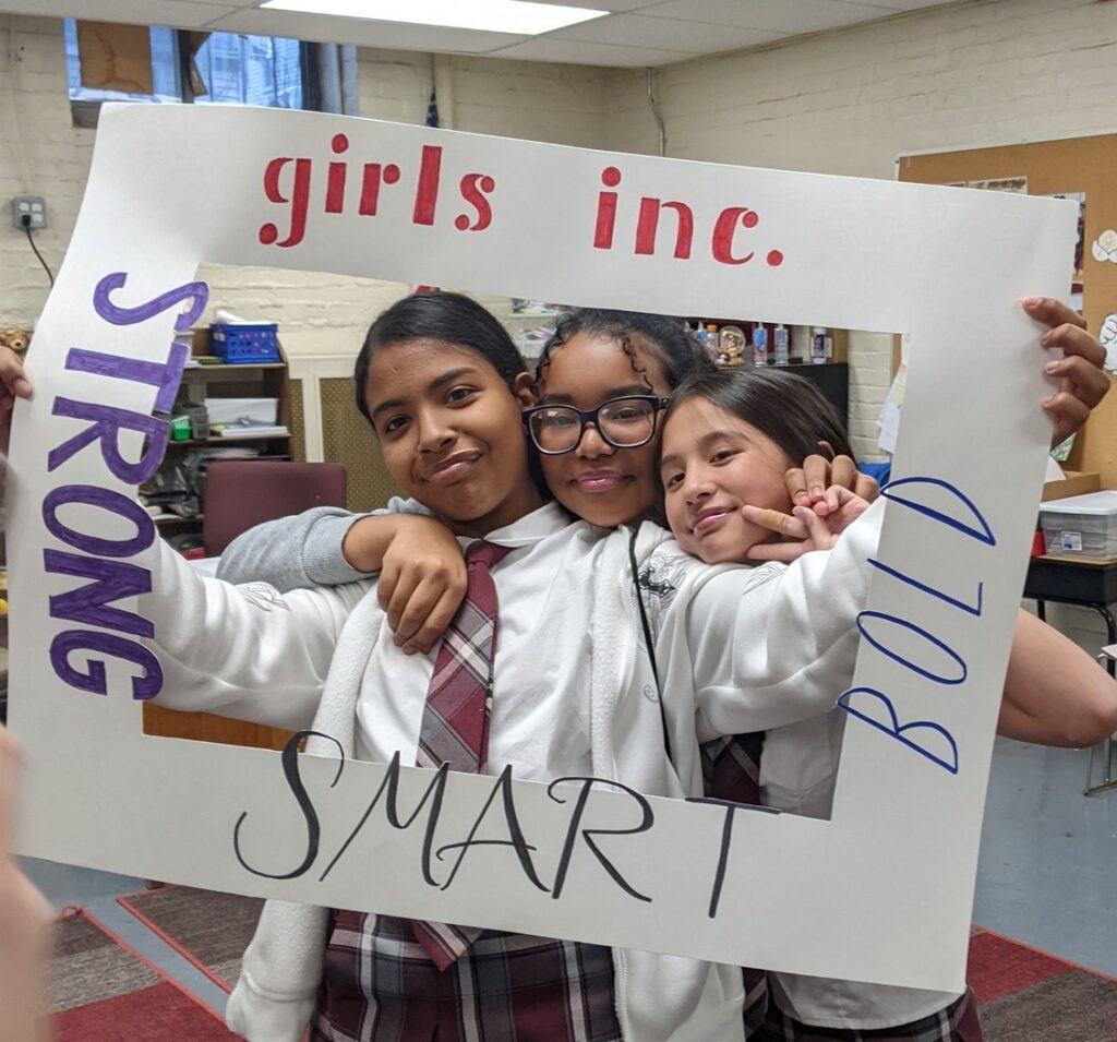 A group of three smiling girls hold a frame around them that reads GIRLS INC. STRONG SMART BOLD