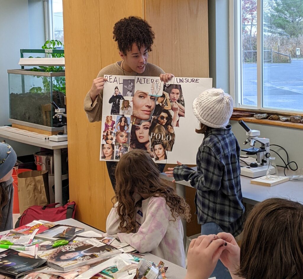 An adult holds up a poster with various magazine cutouts, showing it to a group of young students